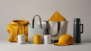 A collection of merchandise is displayed against a neutral background. Visible items include a bright yellow hooded sweatshirt, two canvas tote bags (one light grey and one off-white with dark straps), two white ceramic mugs, one yellow ceramic mug, a yellow baseball cap, and a dark grey thermos with a handle. The items are arranged in a visually appealing composition, suggesting a product showcase or promotional display. apparel