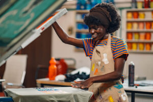 Portrait of smiling multicultural female graphic worker standing near silkscreen printing machine and looking at finished textile product at printing workshop. Females in graphic industry. Copy space.