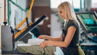 Profile view of a female DTF printing workshop employee standing at facility and using heating press on a silkscreen printed textile product. Side view of a female graphic technician using heat press.